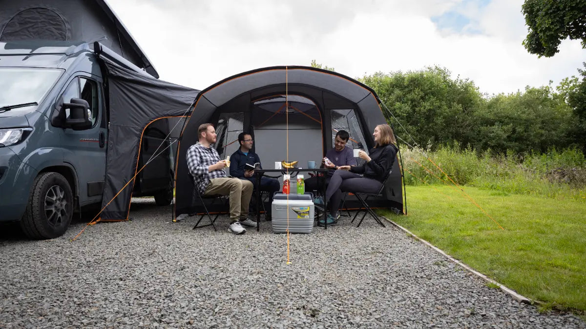 Group of people sitting under the Vango Cove III Mid Poled Awning attached to a van in a natural setting.