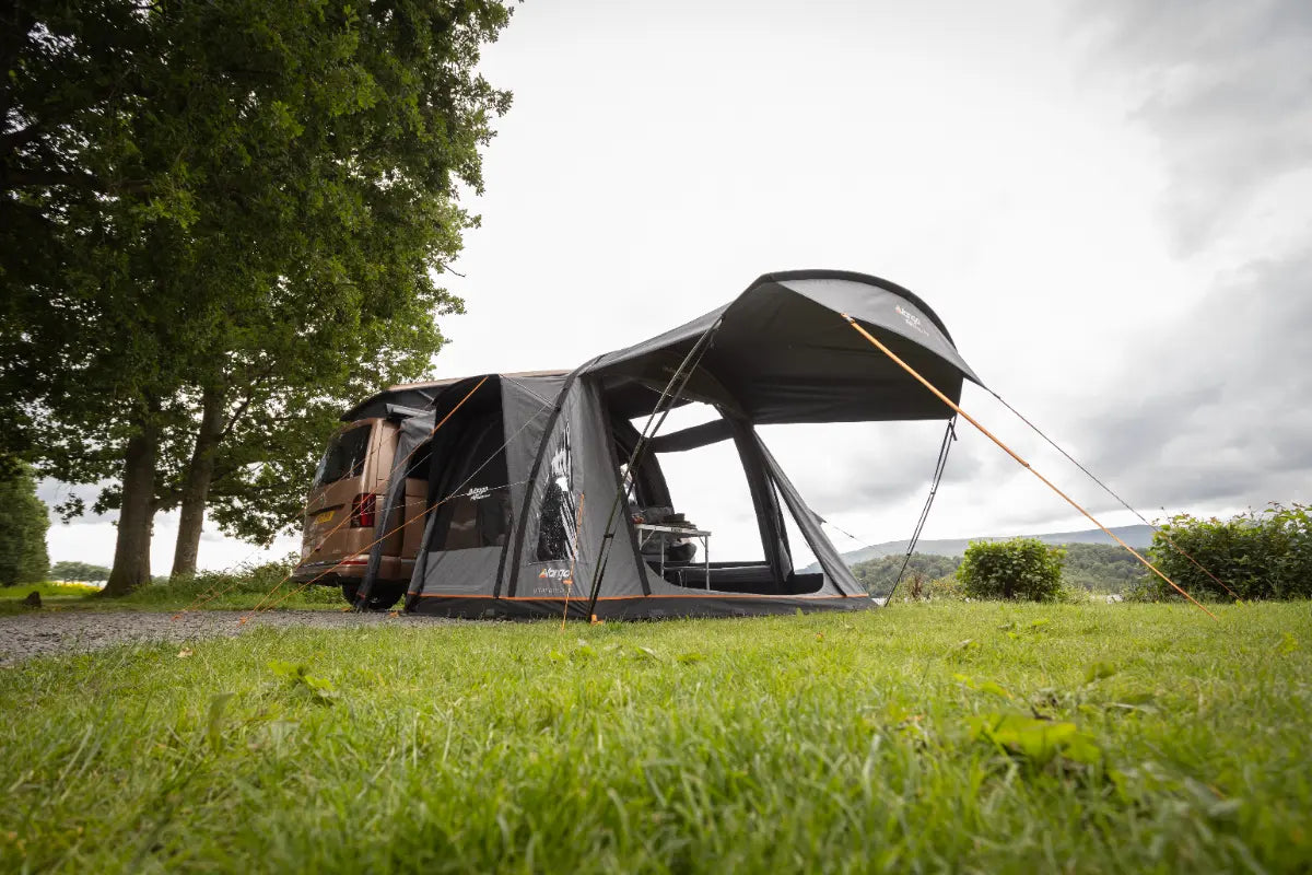 Campervan with an attached Vango Kela Air Low 60th Aniversary Awning on a grassy area with trees and a cloudy sky.