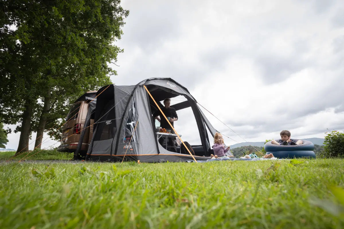 Family enjoying a day outdoors with the Vango Kela Air Low 60th Aniversary Awning set up on grass near a lake.