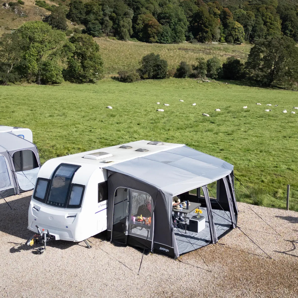 Vango Tuscany Pro Air 500 awning set up at a campsite, with a green field and trees in the background. Another awning is partially visible on the left.