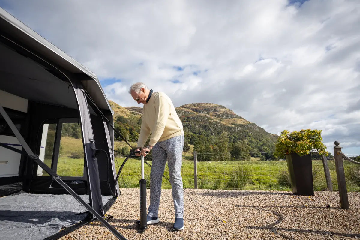 A man inflating the Vango Tuscany Pro Air 500 awning on a gravel campsite with a mountain and cloudy sky in the background.